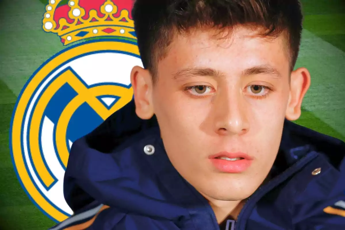 Jove amb jaqueta blava en primer pla amb l’escut del Reial Madrid de fons sobre un camp de futbol