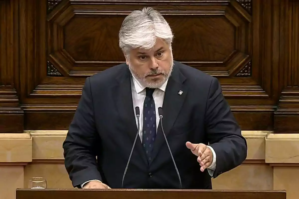 Hombre de cabello canoso y barba hablando en un podio con fondo de madera, vestido con traje oscuro y corbata azul, gesticulando con la mano derecha.