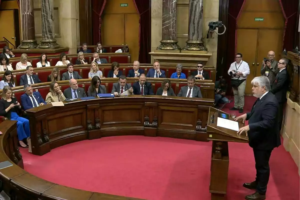 Un hombre de cabello canoso habla desde un atril en una sala parlamentaria mientras varias personas lo escuchan sentadas en bancas de madera y algunos fotógrafos lo observan de pie al fondo