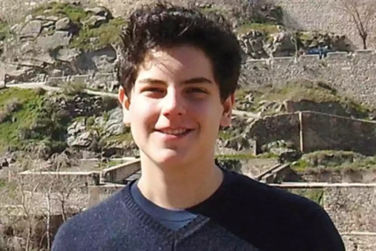 Joven de cabello oscuro sonríe al aire libre con un fondo de paisaje rocoso y vegetación