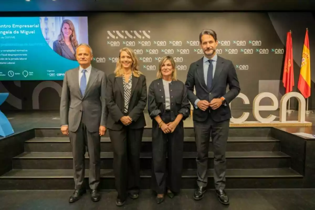 Cuatro personas posan en un escenario de un evento empresarial con fondo negro y logotipos de CEN y dos banderas a la derecha Cuatro personas posan en un escenario de un evento empresarial con fondo negro y logotipos de CEN y dos banderas a la derecha