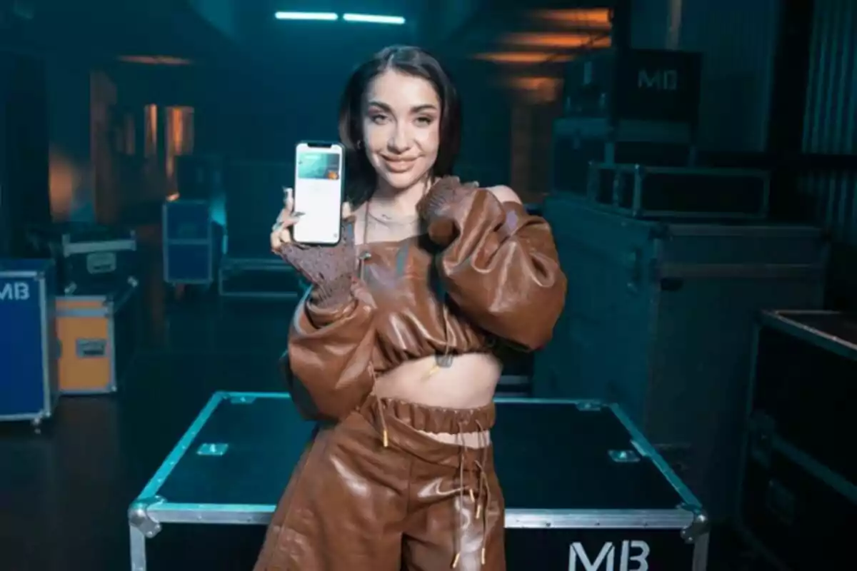 Mujer joven con conjunto marrón sonríe y muestra un teléfono móvil en un ambiente industrial con luces tenues y cajas de equipo en el fondo