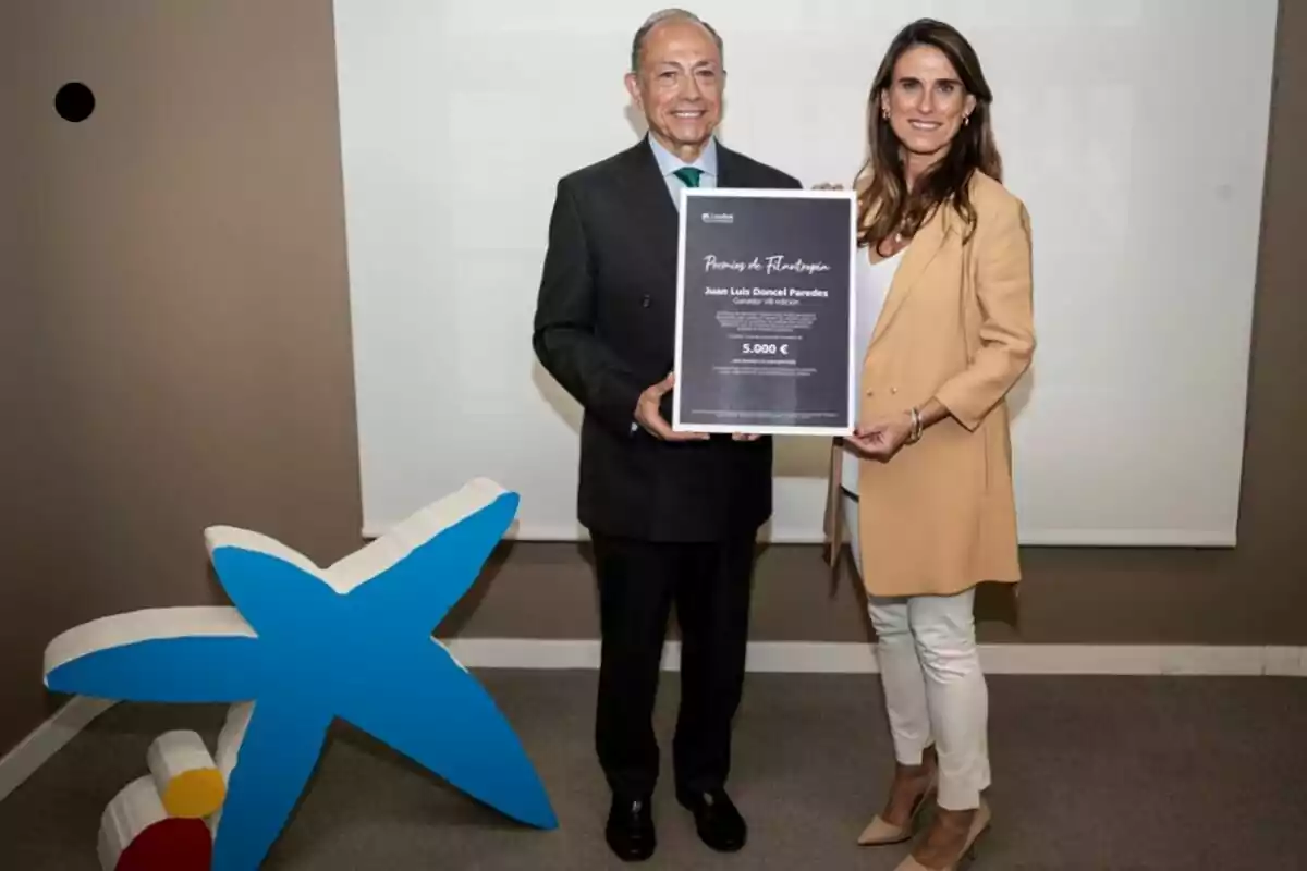 Dos personas posan sonrientes mientras sostienen un diploma de reconocimiento, junto a una figura decorativa azul en forma de estrella.