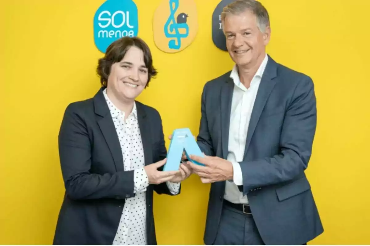 Dues persones somrients amb vestits formals sostenen un trofeu blau davant d’una paret groga amb logotips de fons Dues persones somrients amb vestits formals sostenen un trofeu blau davant d’una paret groga amb logotips de fons