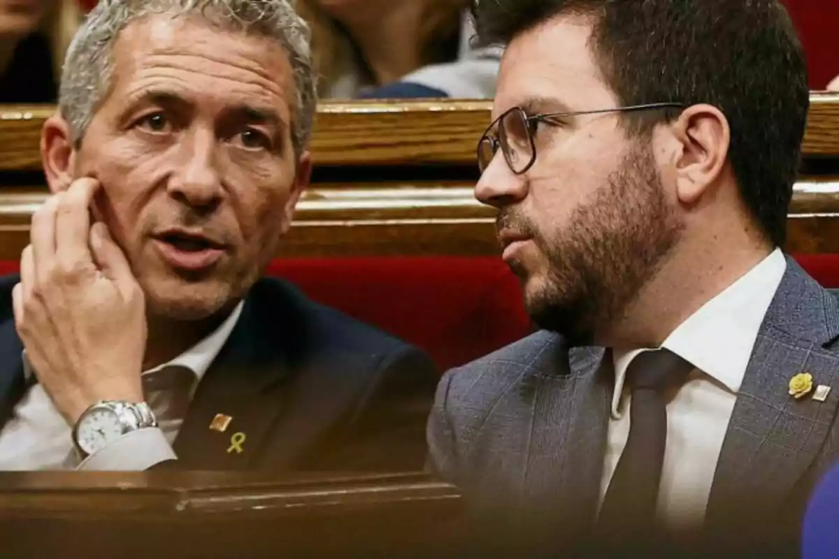 Dos hombres con traje conversan sentados en un banco de madera en un entorno formal