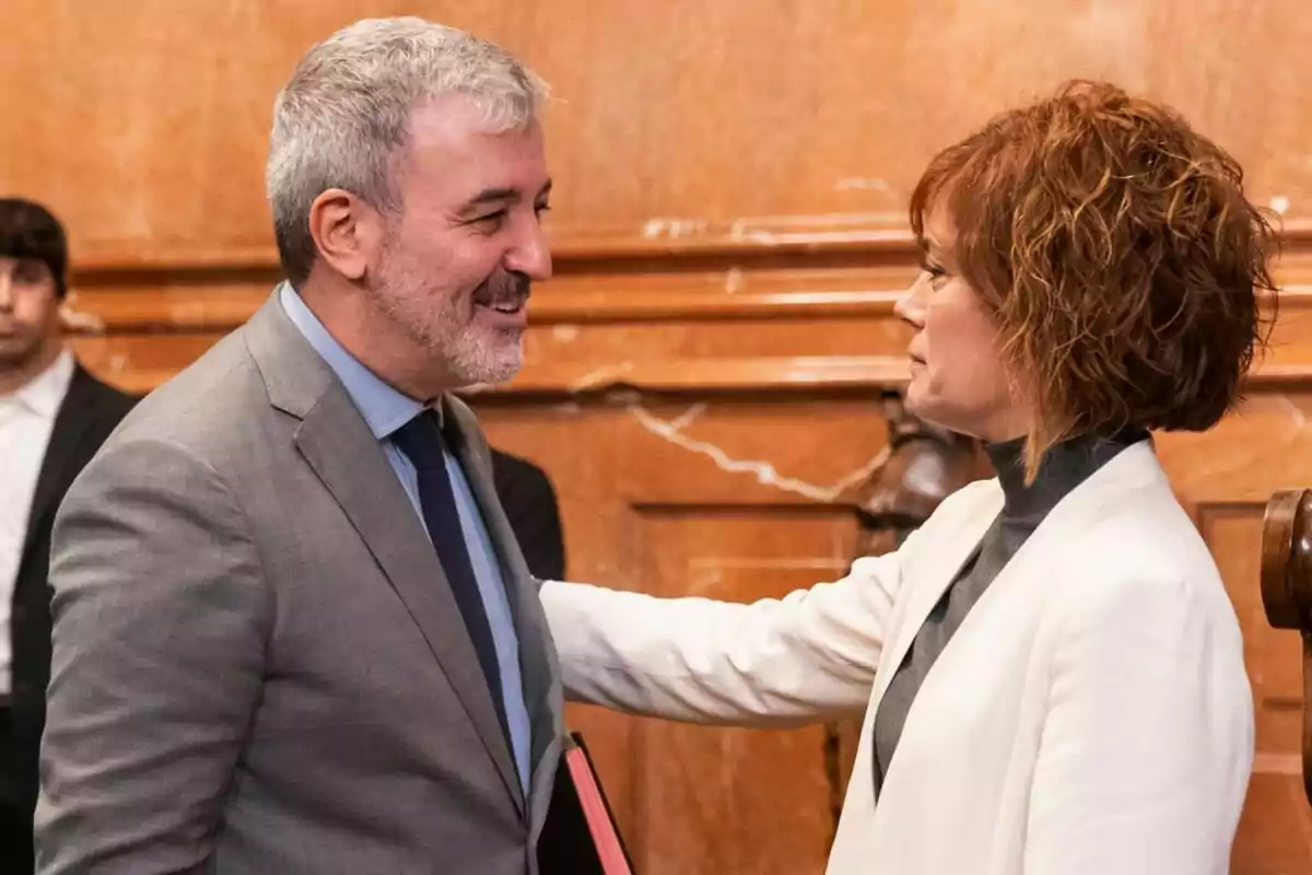 Un hombre de cabello canoso y barba sonríe mientras una mujer de cabello corto y rojizo lo saluda con una mano en el hombro, ambos vestidos de manera formal en un entorno interior elegante.