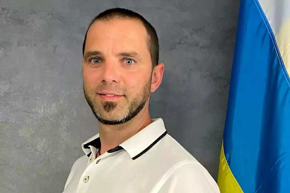 Hombre de cabello corto y barba recortada viste camisa blanca y está de pie junto a una bandera azul y amarilla sobre fondo gris