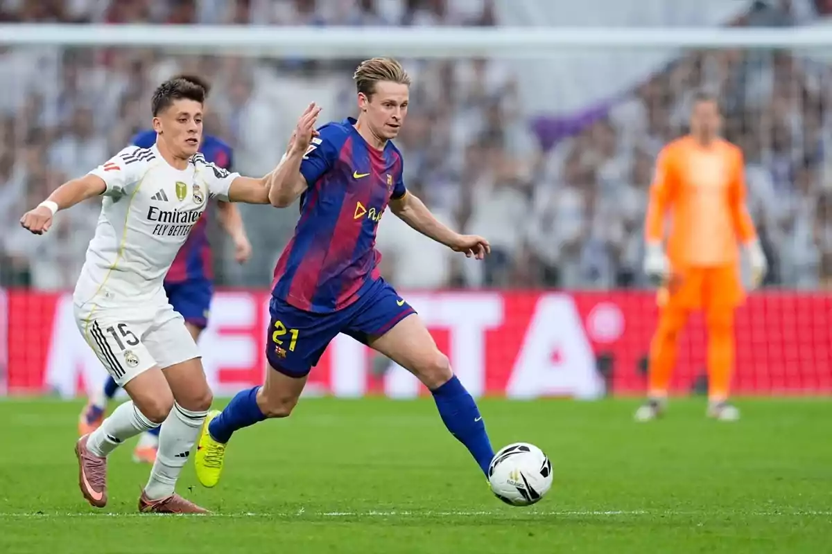 Dos futbolistes dels equips Reial Madrid i Barcelona disputen la pilota durant un partit mentre el porter observa des del fons. Dos futbolistes dels equips Reial Madrid i Barcelona disputen la pilota durant un partit mentre el porter observa des del fons.