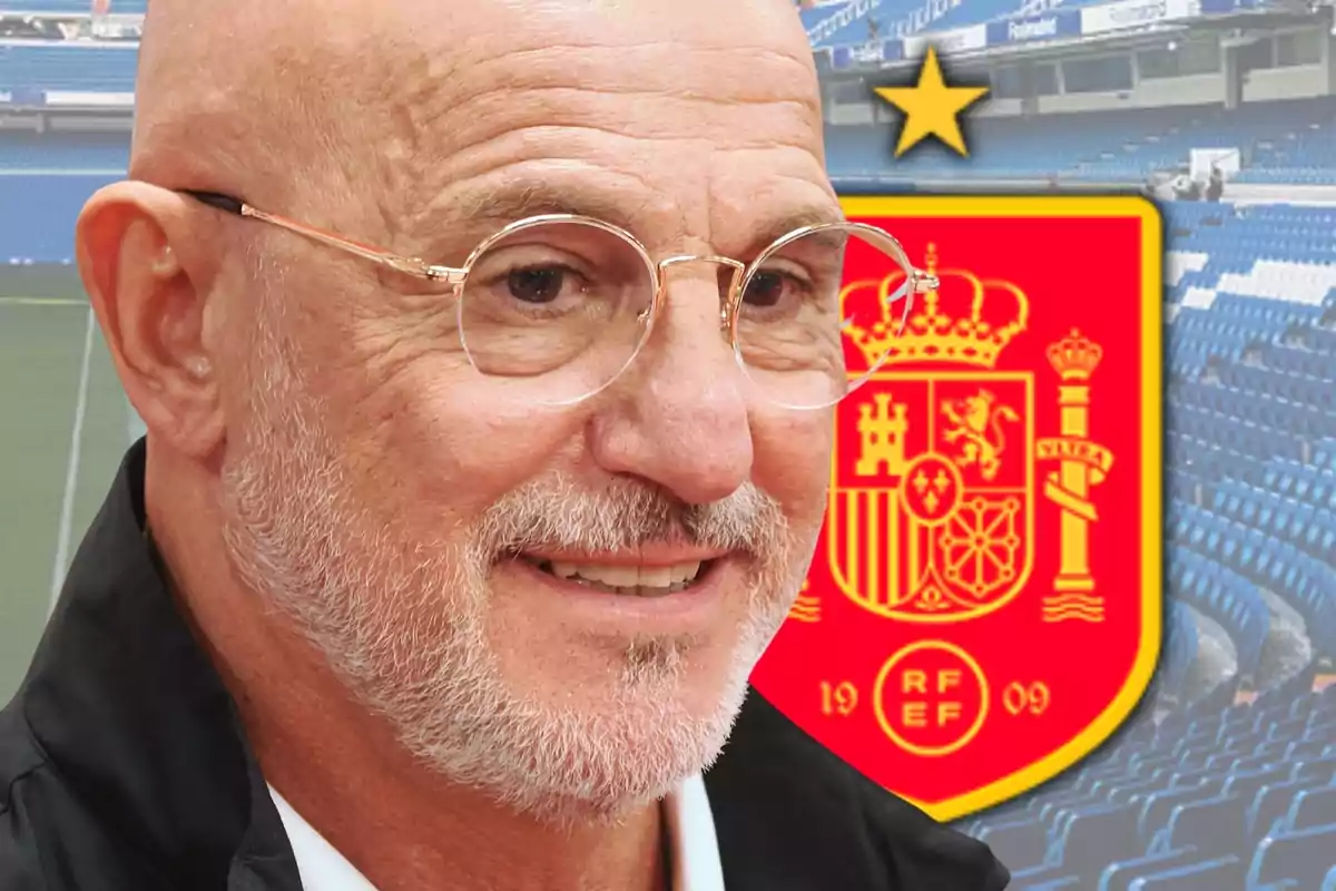 Home calb amb ulleres i barba somriu davant l’escut de la selecció espanyola de futbol en un estadi.