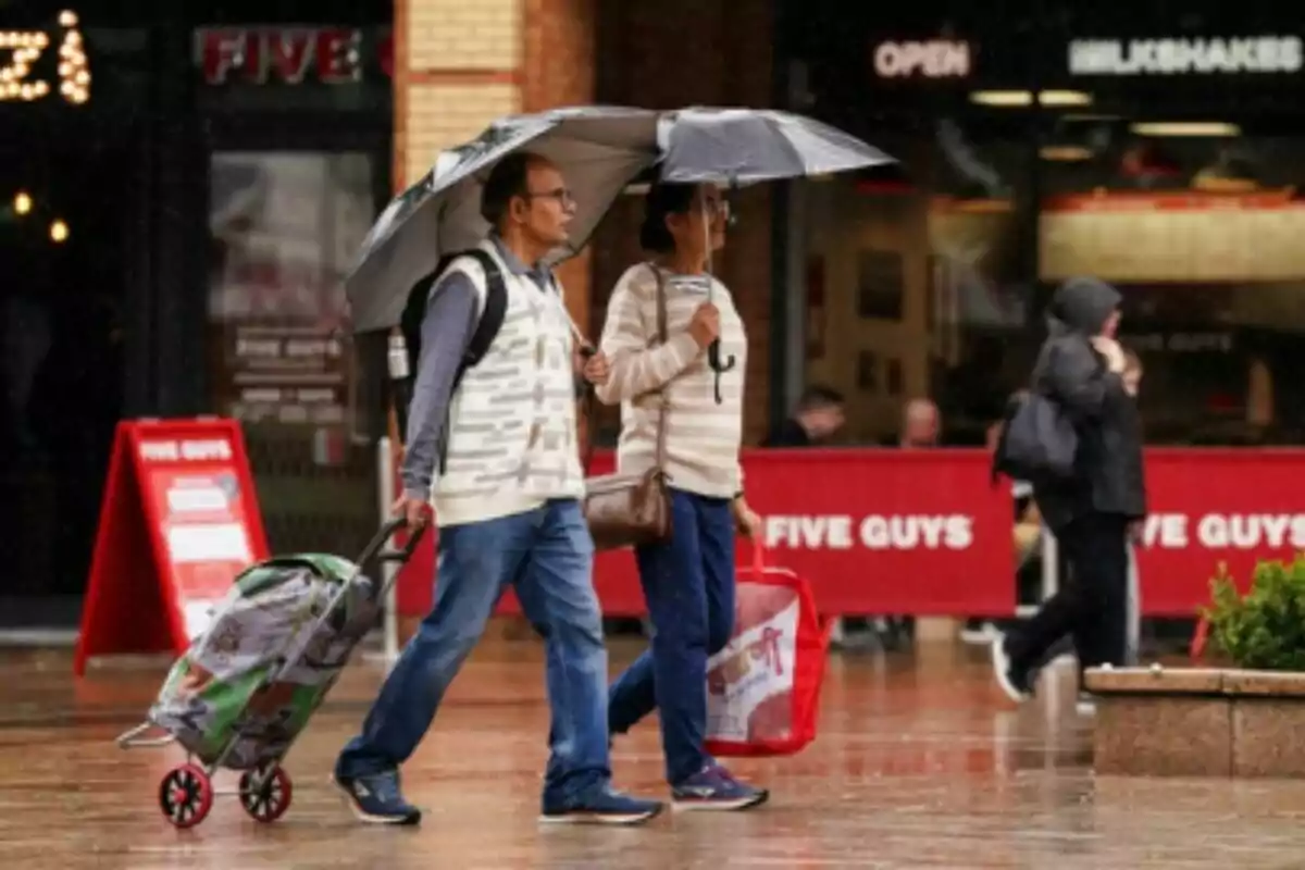 Dos personas caminan bajo la lluvia usando paraguas y llevando bolsas de compras frente a un restaurante Five Guys.