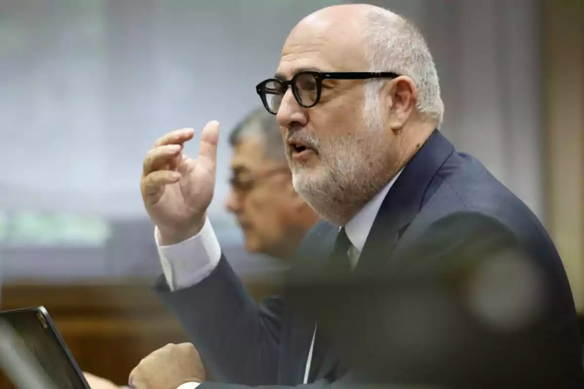 Hombre de mediana edad con barba y gafas hablando en una reunión formal mientras gesticula con la mano