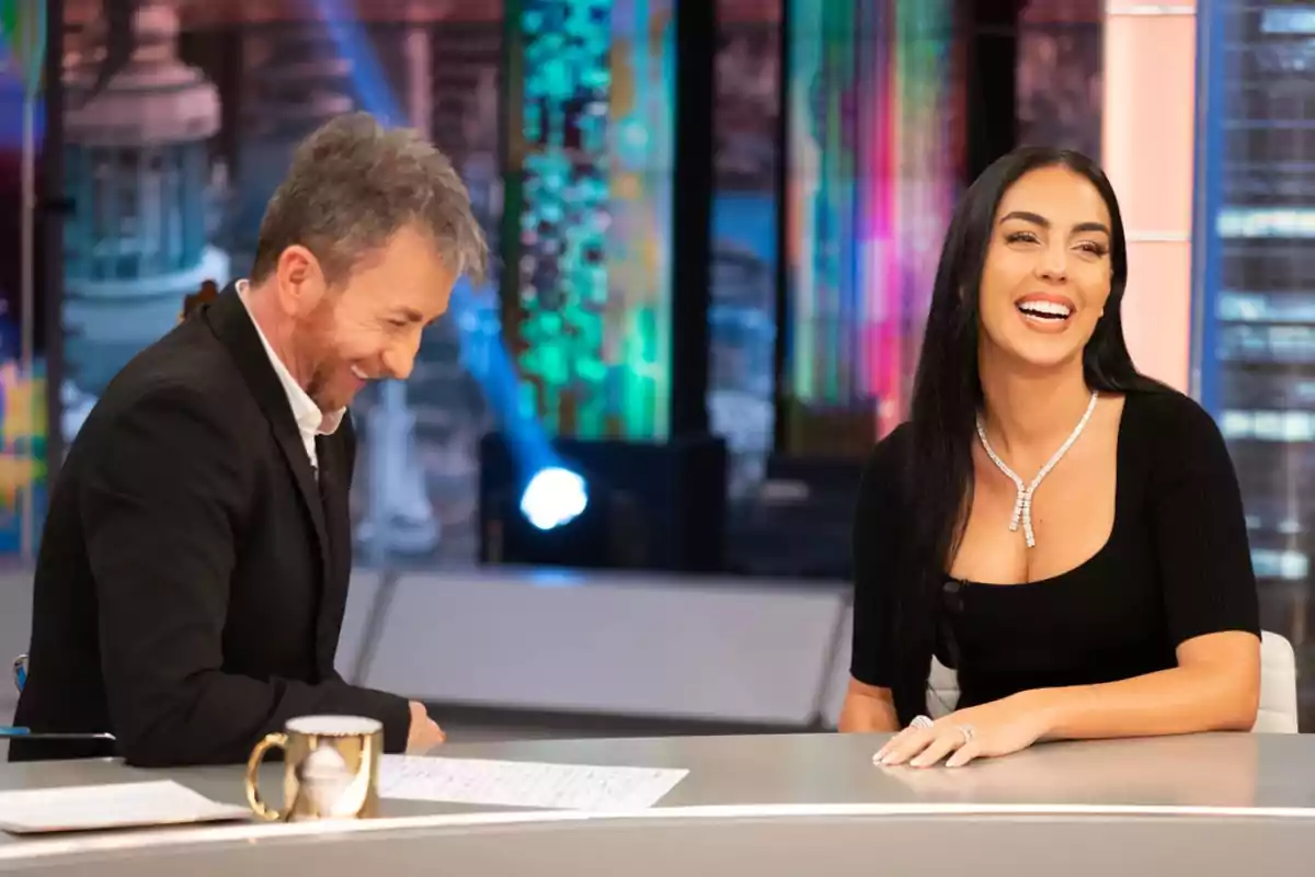 Pablo Motos y Georgina Rodríguez sentadas en un set de televisión conversan y sonríen mientras están frente a una mesa con una taza y papeles. Captura de 'El Hormiguero'