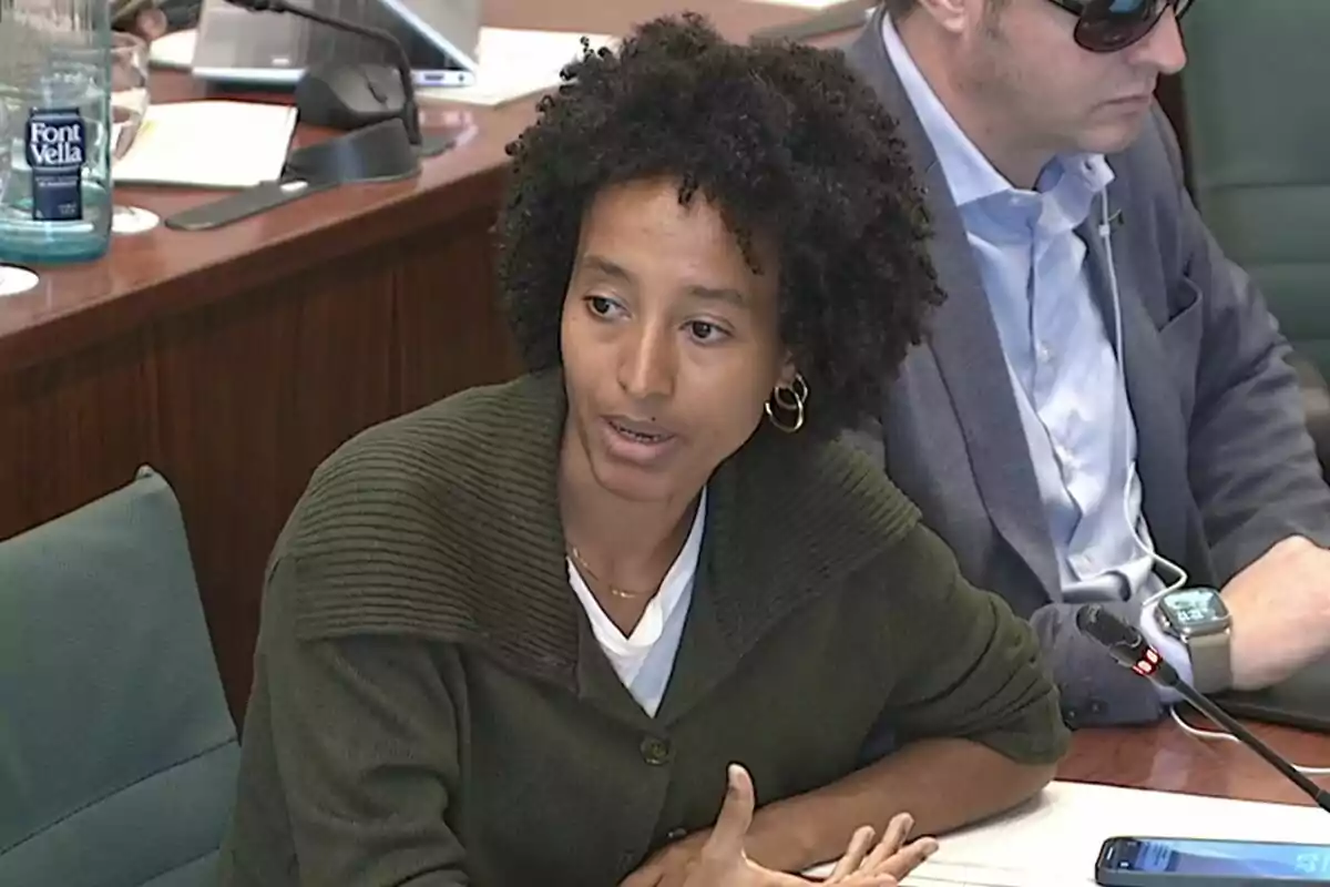 Mujer hablando en una reunión mientras un hombre con gafas de sol está sentado a su lado