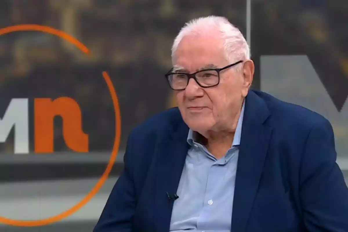 Hombre mayor de cabello canoso y gafas con saco azul sentado en un estudio de televisión