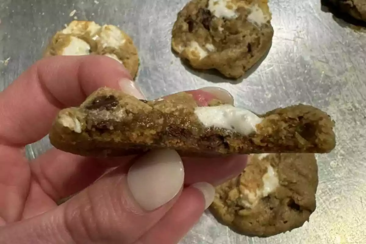 Galleta partida a la mitad mostrando su interior con trozos de chocolate y malvavisco de Target, sostenida por una mano sobre una bandeja metálica. Galleta partida a la mitad mostrando su interior con trozos de chocolate y malvavisco de Target, sostenida por una mano sobre una bandeja metálica.