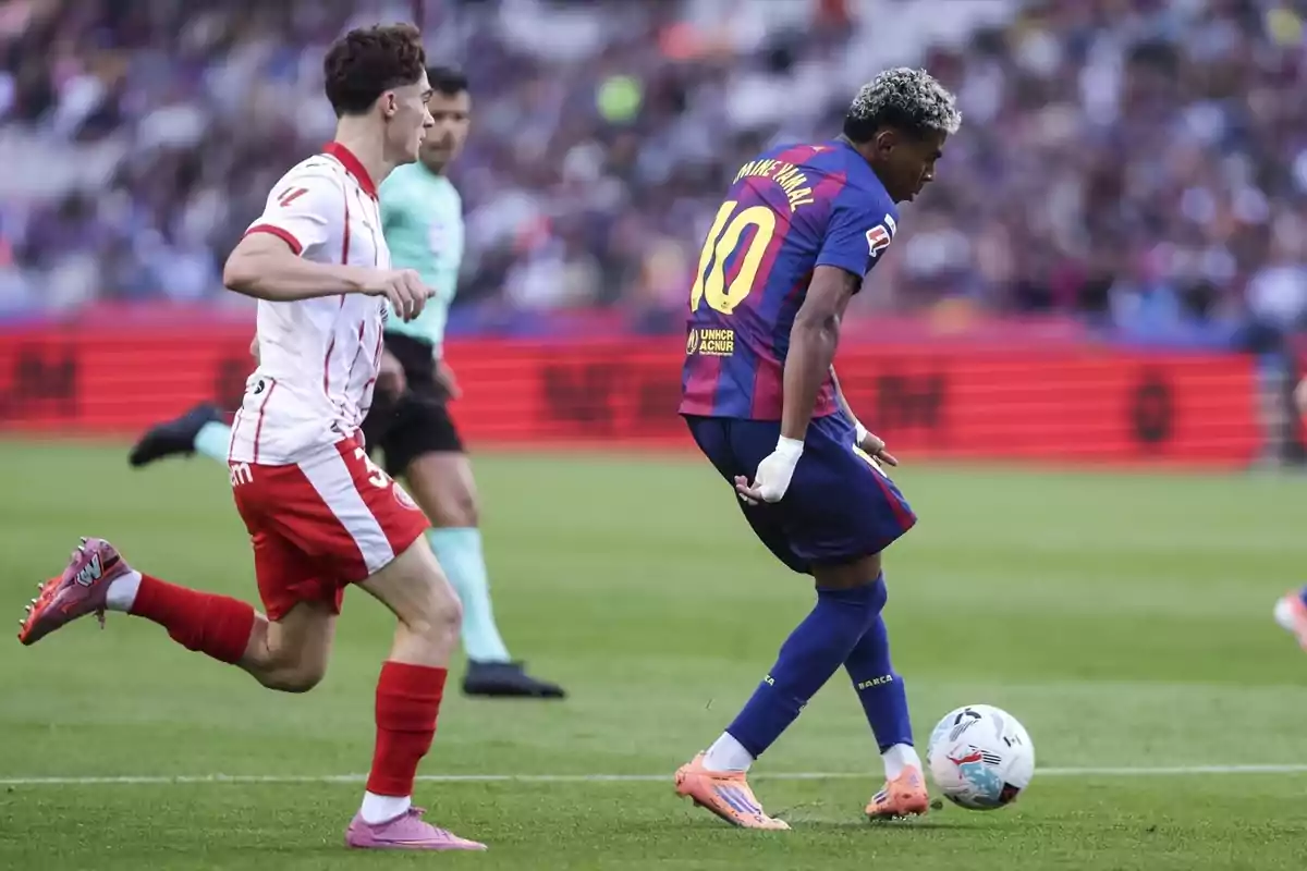 Dos futbolistes disputen la pilota durant un partit, un amb uniforme blaugrana i l’altre amb uniforme blanc i vermell, mentre un àrbitre observa la jugada al fons. Dos futbolistes disputen la pilota durant un partit, un amb uniforme blaugrana i l’altre amb uniforme blanc i vermell, mentre un àrbitre observa la jugada al fons.