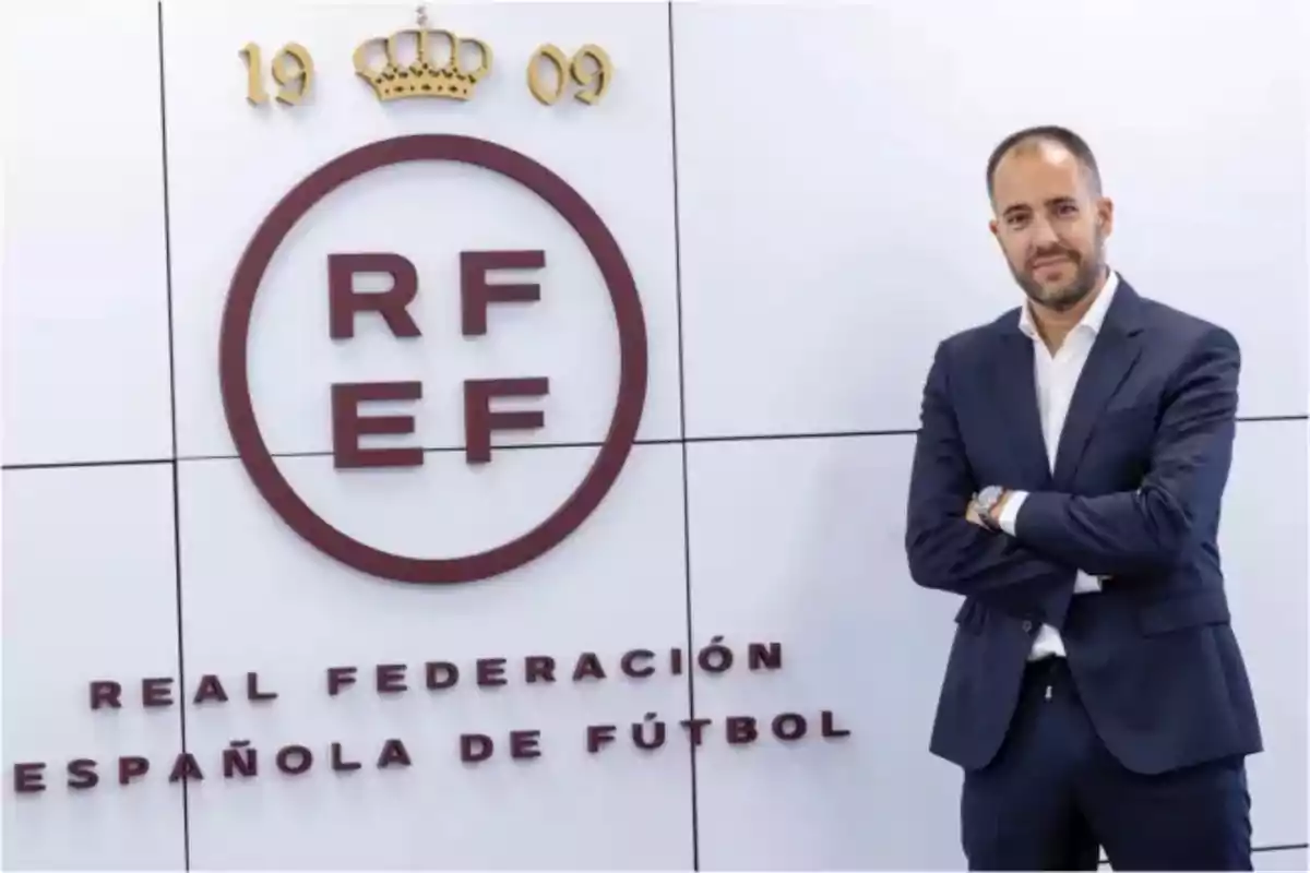 Home amb vestit posant al costat del logotip de la Reial Federació Espanyola de Futbol en una paret blanca