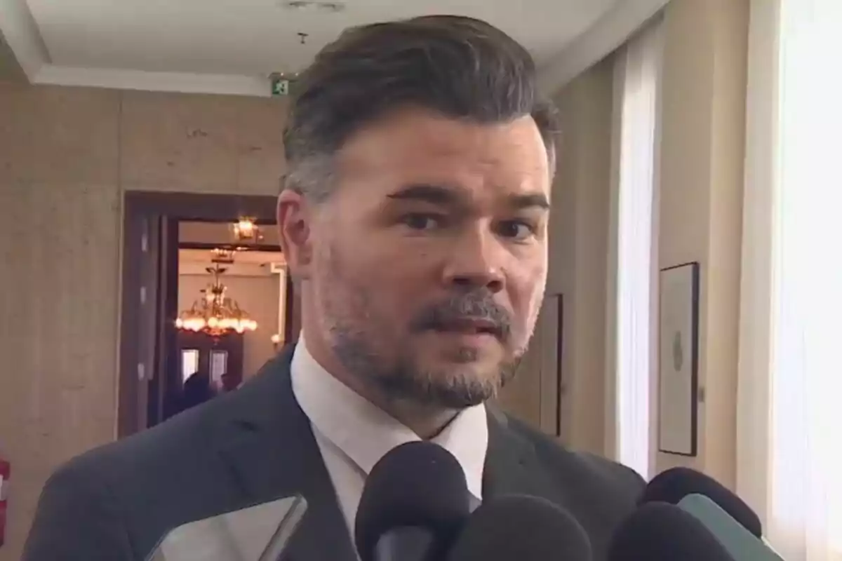 Hombre de cabello oscuro y barba corta vestido con traje y corbata hablando frente a varios micrófonos en un interior elegante con lámparas y paredes claras
