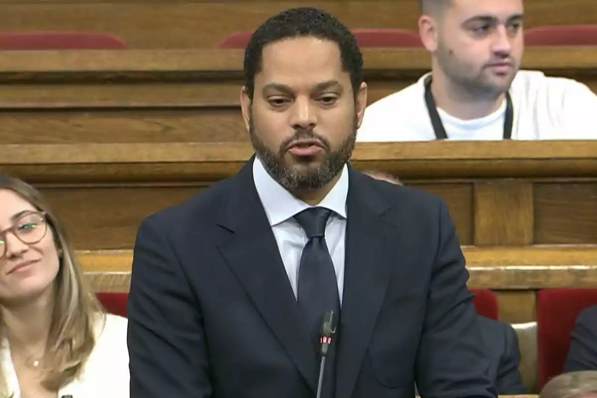 Hombre con barba y traje oscuro hablando frente a un micrófono en un recinto con bancas de madera y otras personas sentadas al fondo