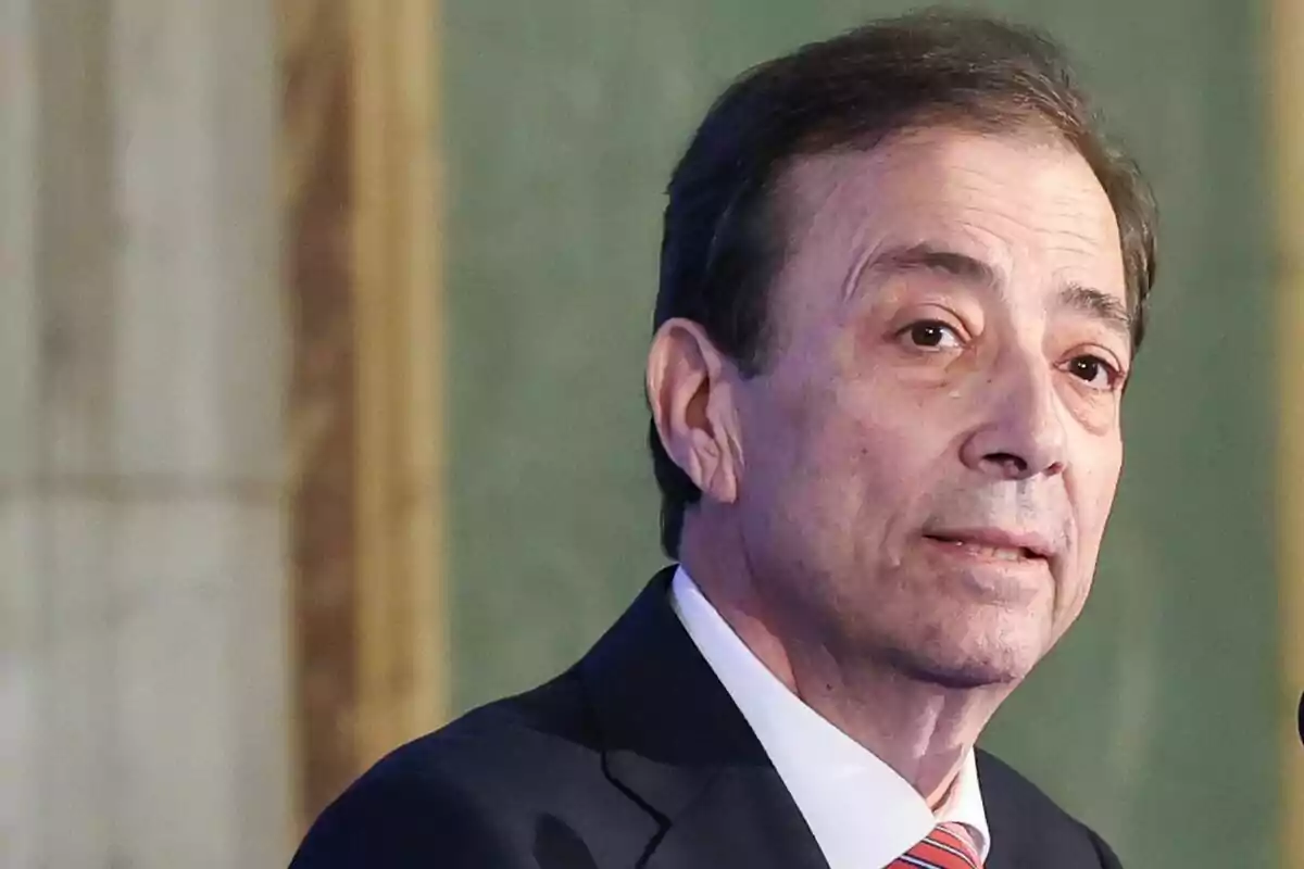 Home de mitjana edat amb cabell fosc i vestit formal parlant davant d’un micròfon en un entorn interior elegant