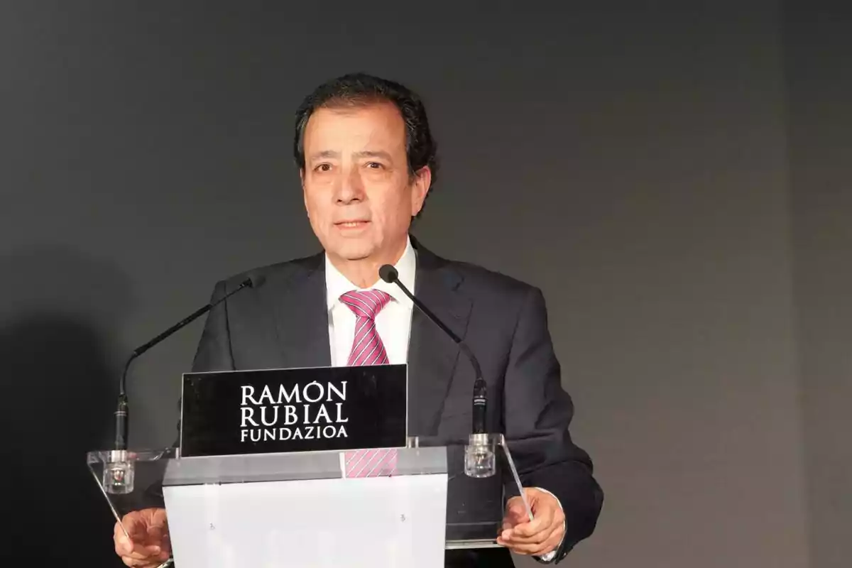 Home amb vestit fosc i corbata rosa parlant en un faristol amb el rètol Ramón Rubial Fundazioa sobre fons gris