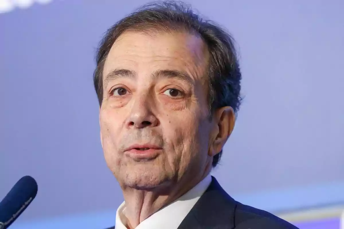 Hombre de mediana edad con cabello oscuro y canoso usando traje oscuro y camisa blanca hablando frente a un micrófono con fondo azul claro