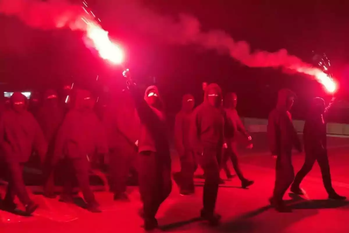 Grupo de personas encapuchadas caminando de noche mientras sostienen bengalas rojas que iluminan la escena