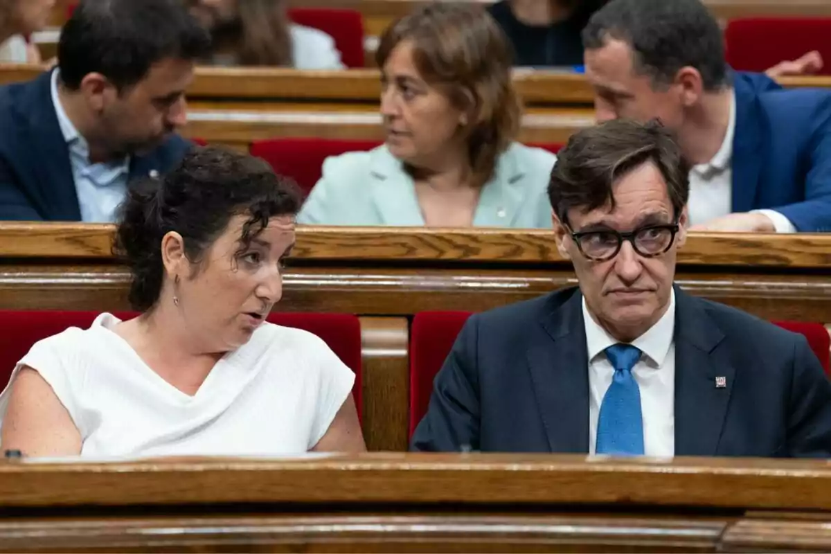 Dues persones assegudes en un banc vermell conversen en el que sembla ser un parlament mentre altres persones estan assegudes darrere d’elles.
