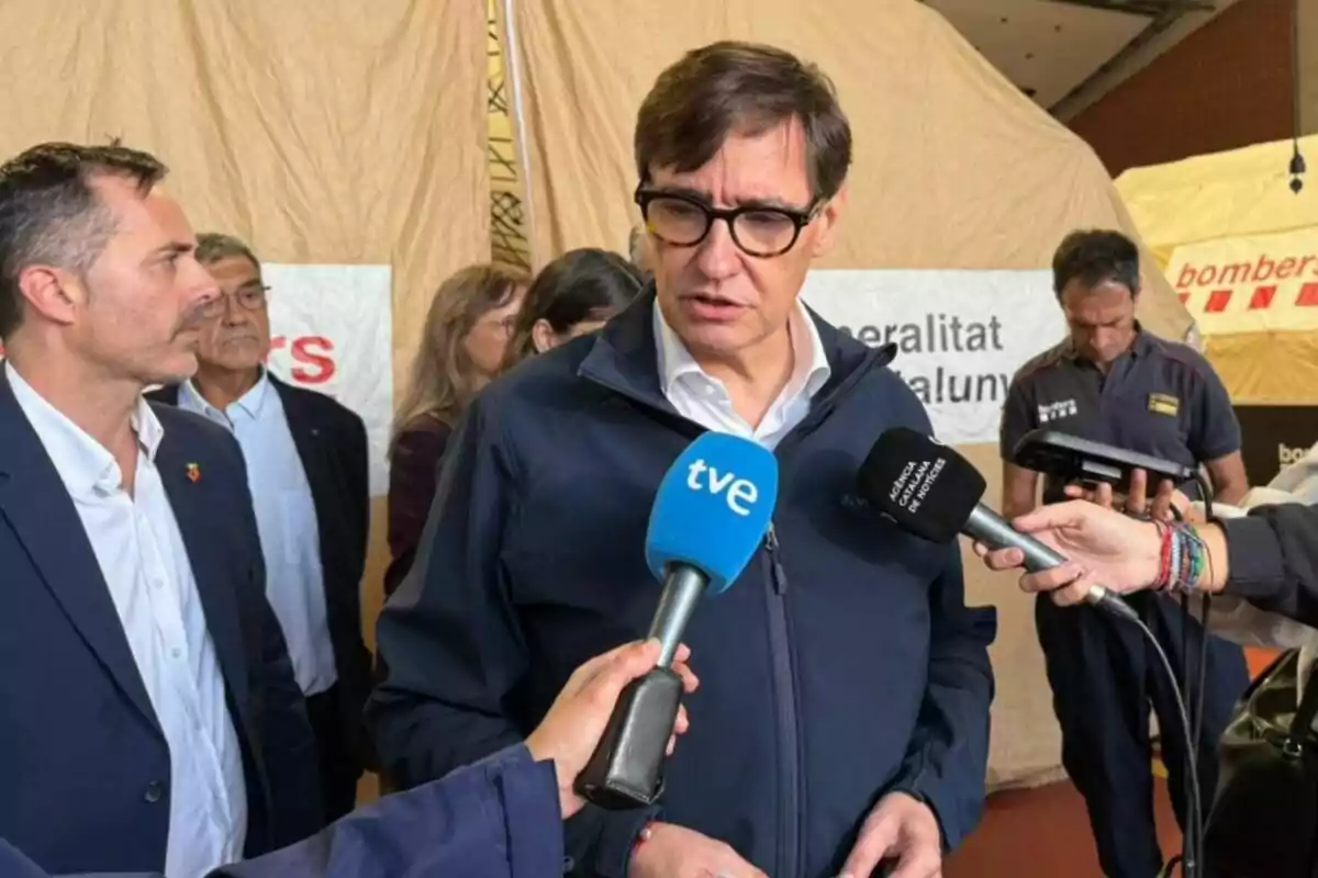 Un hombre con gafas y chaqueta azul responde preguntas de varios periodistas que sostienen micrófonos frente a él, mientras otras personas observan en el fondo y se ven carpas de emergencia.