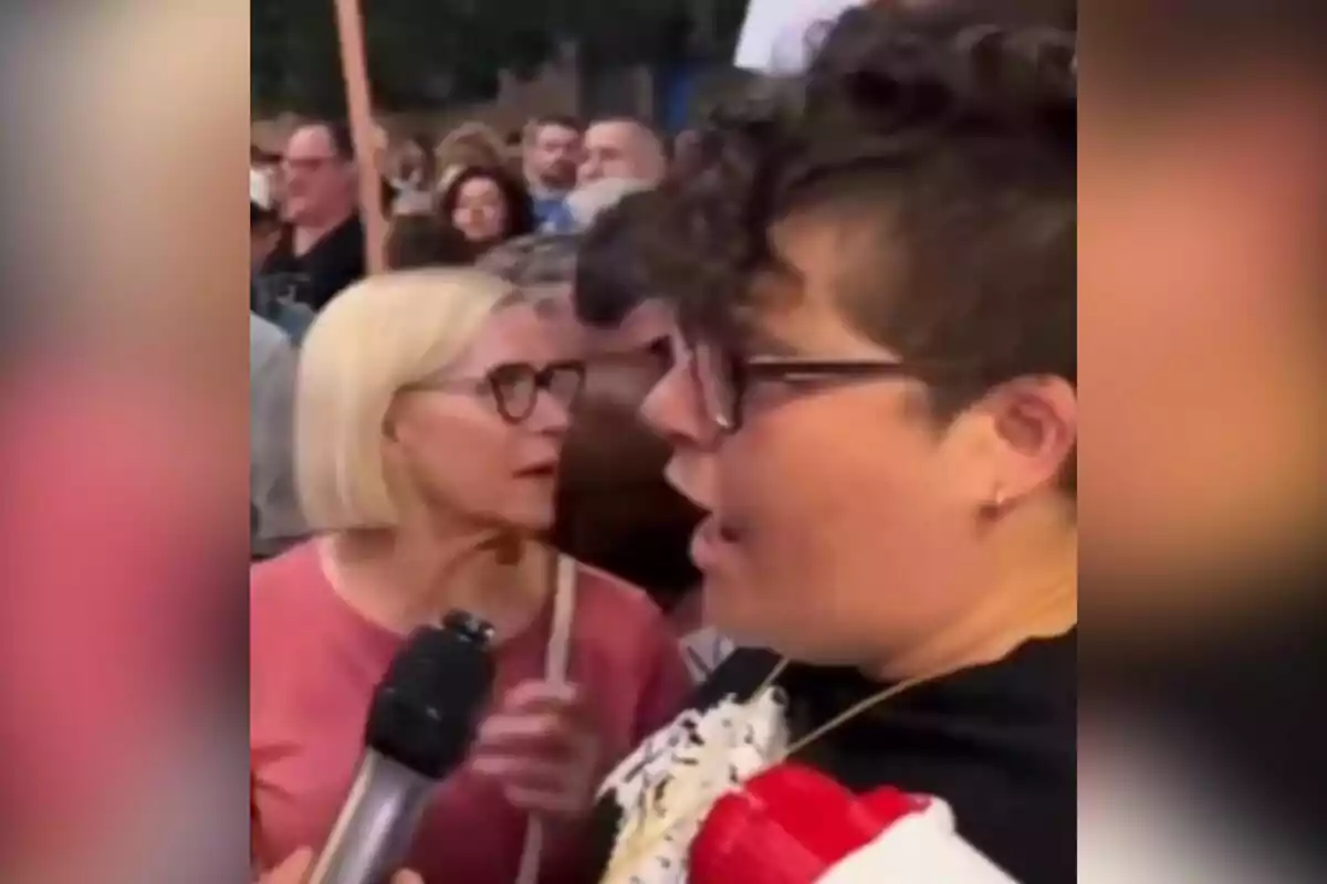 Dos personas con gafas conversan en medio de una multitud mientras una de ellas sostiene un micrófono.