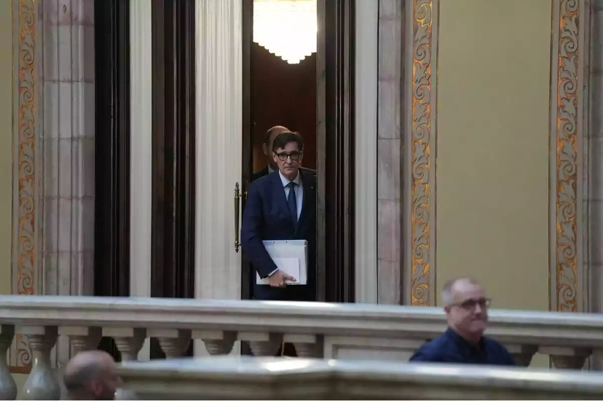Hombre con gafas y traje azul sale de una puerta sosteniendo documentos en un edificio con detalles arquitectónicos ornamentados