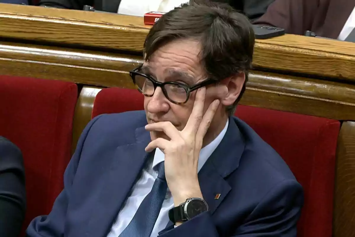 Hombre con gafas y traje azul sentado en un banco rojo, apoyando la cabeza en su mano y mirando pensativo hacia un lado Hombre con gafas y traje azul sentado en un banco rojo, apoyando la cabeza en su mano y mirando pensativo hacia un lado