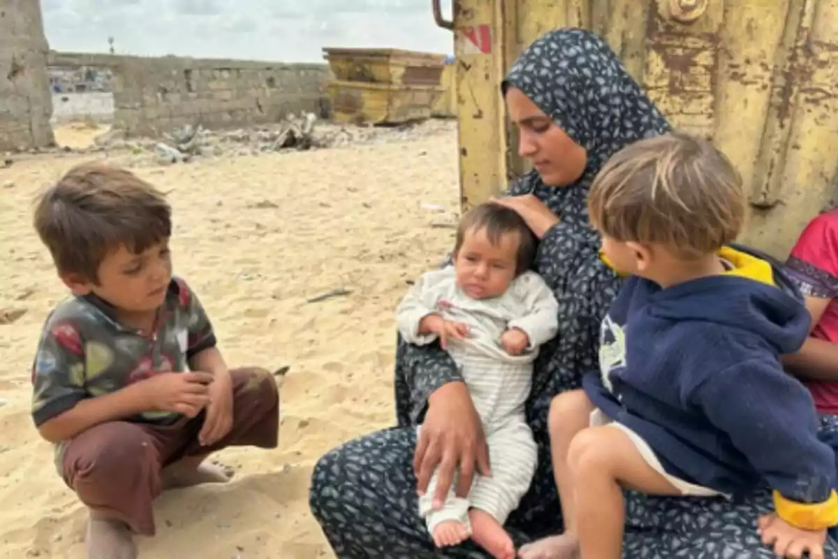 Dona asseguda a la sorra sostenint un nadó en braços, acompanyada per dos nens petits, amb un fons d’estructures deteriorades després del conflicte a Gaza. Dona asseguda a la sorra sostenint un nadó en braços, acompanyada per dos nens petits, amb un fons d’estructures deteriorades després del conflicte a Gaza.