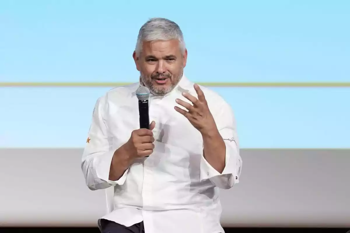 Home de cabells canosos i barba curta vestit amb jaqueta de xef blanca parlant mentre sosté un micròfon en una mà i gesticula amb l’altra, assegut davant d’un fons clar