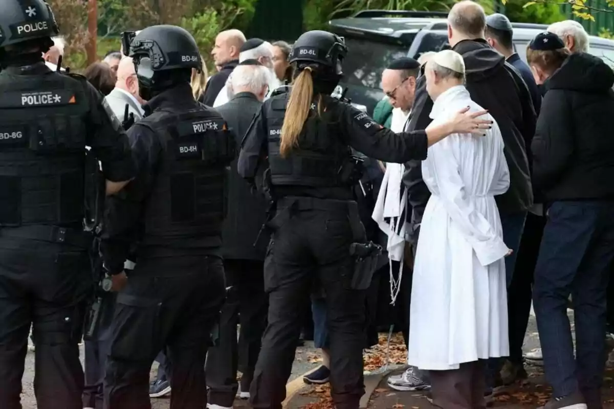 Policies fortament armats custodien un grup de persones vestides amb roba tradicional i religiosa en un carrer envoltat d’arbres i fulles caigudes. Policies fortament armats custodien un grup de persones vestides amb roba tradicional i religiosa en un carrer envoltat d’arbres i fulles caigudes.