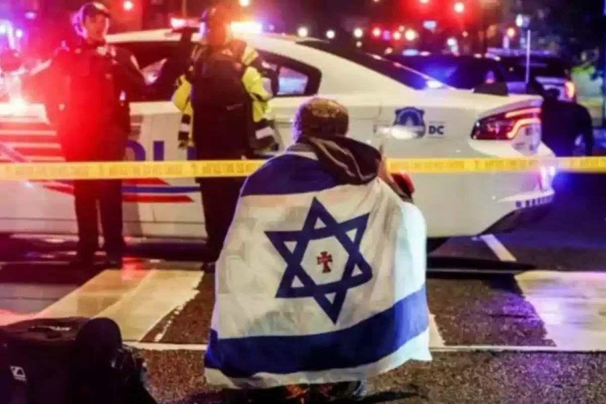 Persona embolicada amb una bandera d’Israel asseguda davant d’una patrulla de policia i cinta de precinte mentre dos agents observen en un carrer il·luminat per llums d’emergència. Persona embolicada amb una bandera d’Israel asseguda davant d’una patrulla de policia i cinta de precinte mentre dos agents observen en un carrer il·luminat per llums d’emergència.
