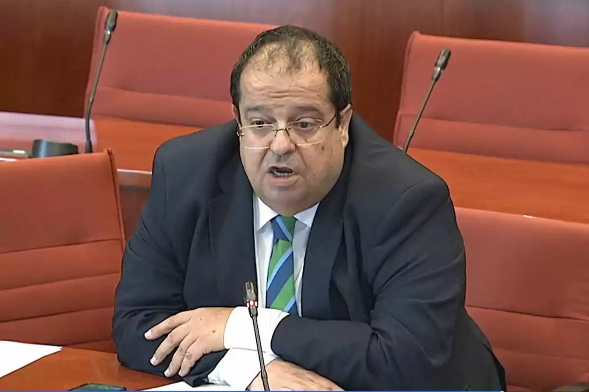 Hombre con gafas y corbata hablando en una sala de reuniones con sillas rojas y micrófonos sobre la mesa