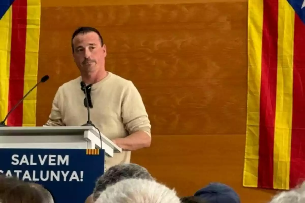 Hombre de pie hablando en un podio con un cartel que dice Salvem Catalunya y dos banderas catalanas a los lados