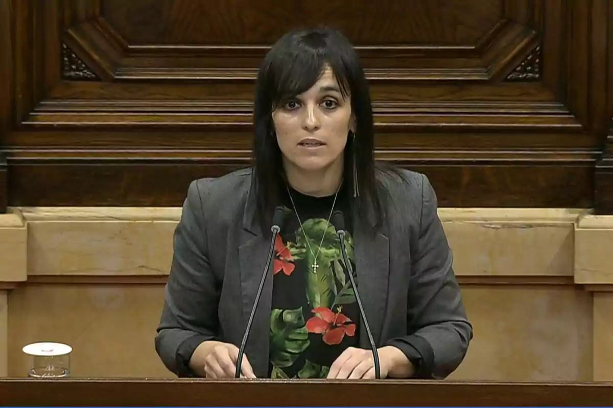 Mujer de cabello oscuro y liso hablando en un atril de madera con dos micrófonos, vestida con chaqueta gris y camiseta negra con estampado de flores rojas y hojas verdes, fondo de madera tallada