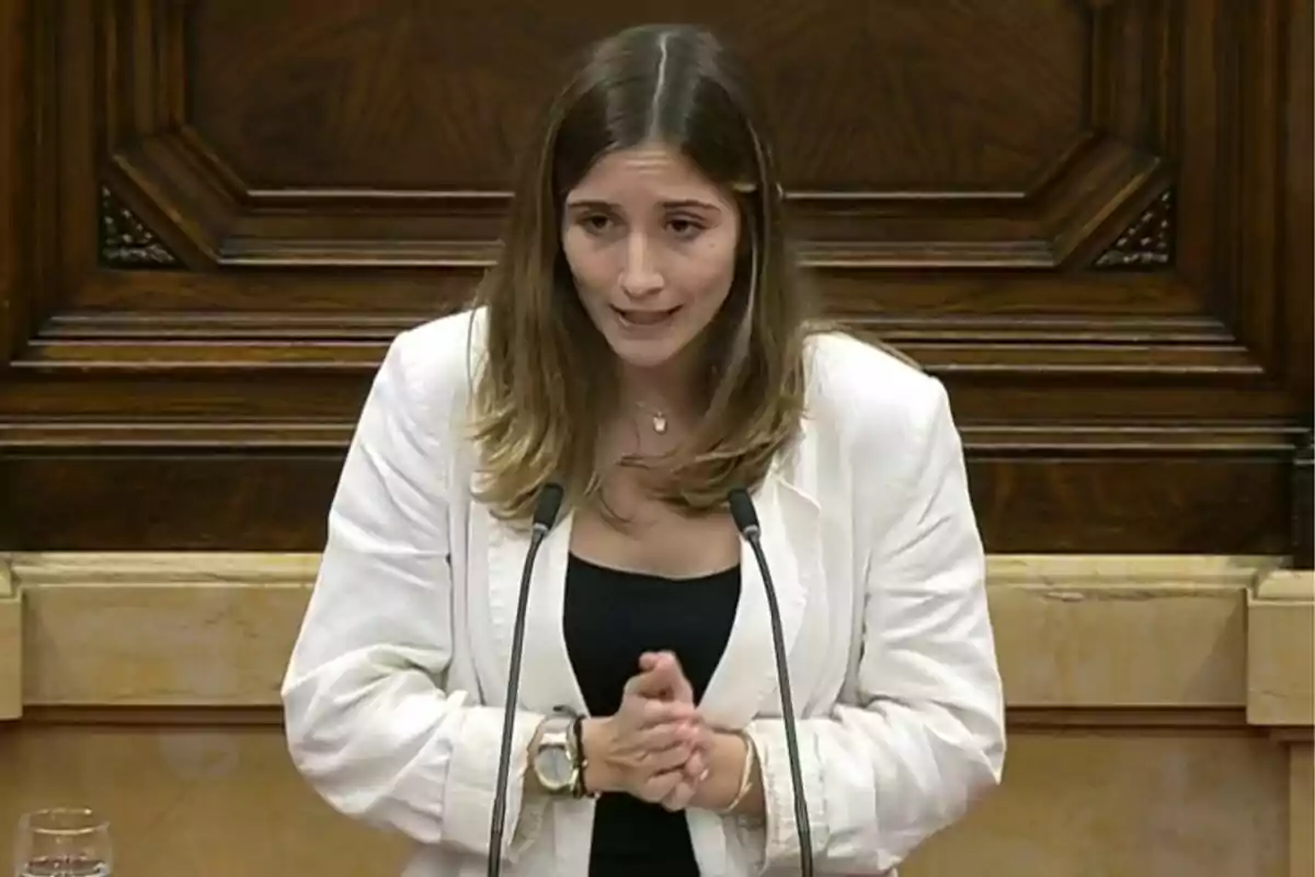 Mujer joven con chaqueta blanca hablando frente a dos micrófonos en un recinto de madera