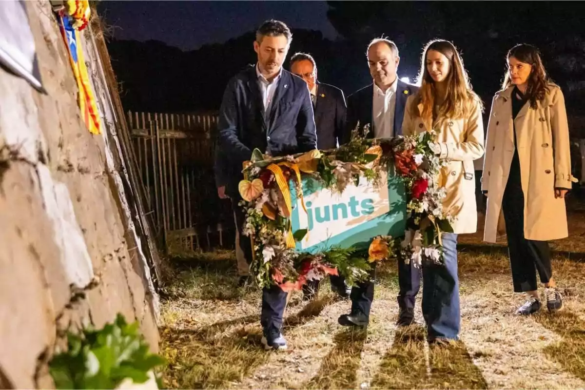 Cinco personas participan en un acto nocturno al aire libre, sosteniendo una corona de flores con la palabra junts, junto a una pared de piedra decorada con una bandera catalana.