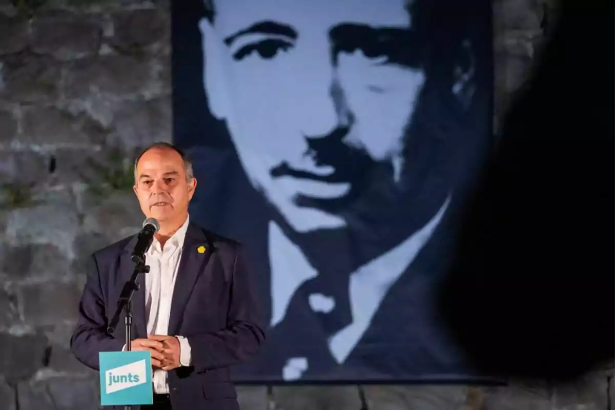 Hombre de traje hablando en un micrófono con un cartel de junts y un retrato grande en blanco y negro de fondo