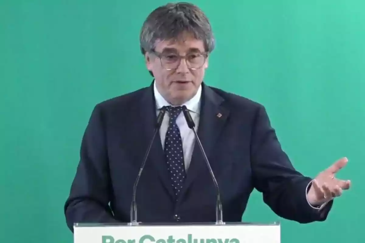 Home de cabells canosos i ulleres parlant en un faristol amb fons verd i micròfons davant seu Home de cabells canosos i ulleres parlant en un faristol amb fons verd i micròfons davant seu