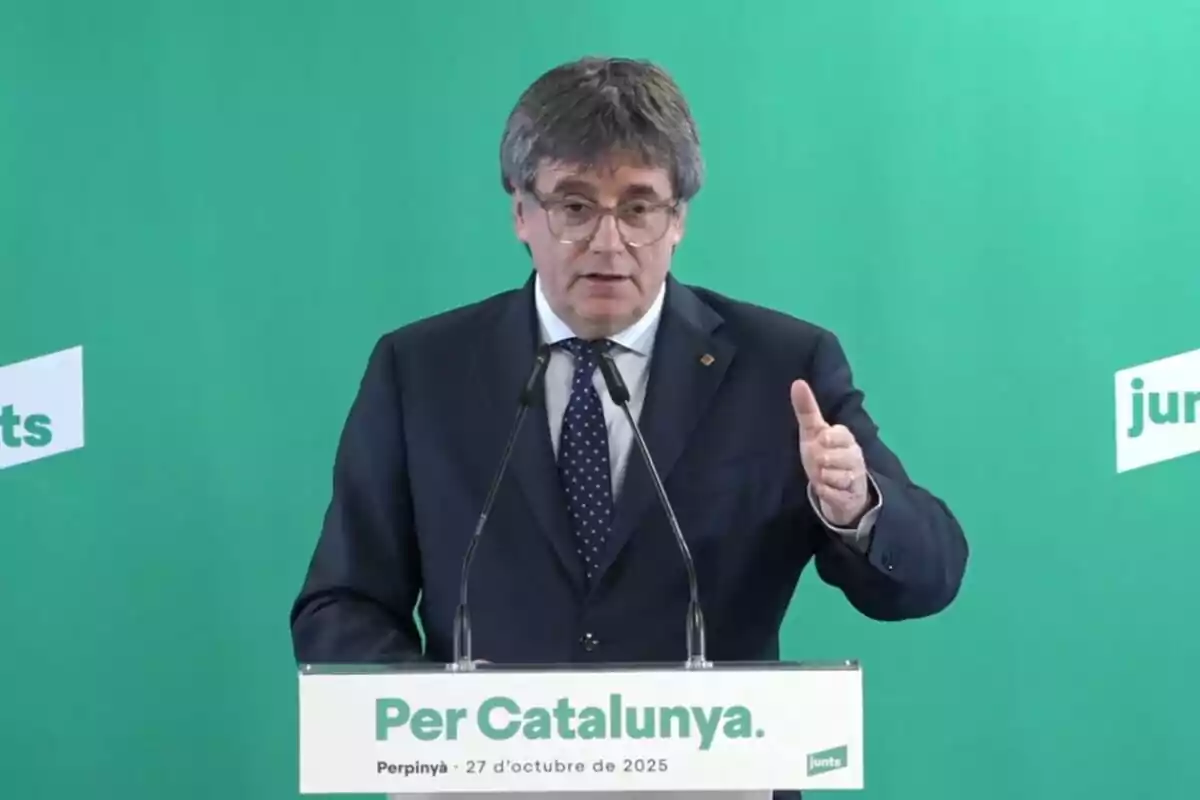 Home amb ulleres i vestit fosc parlant en un faristol amb fons verd i el text Per Catalunya