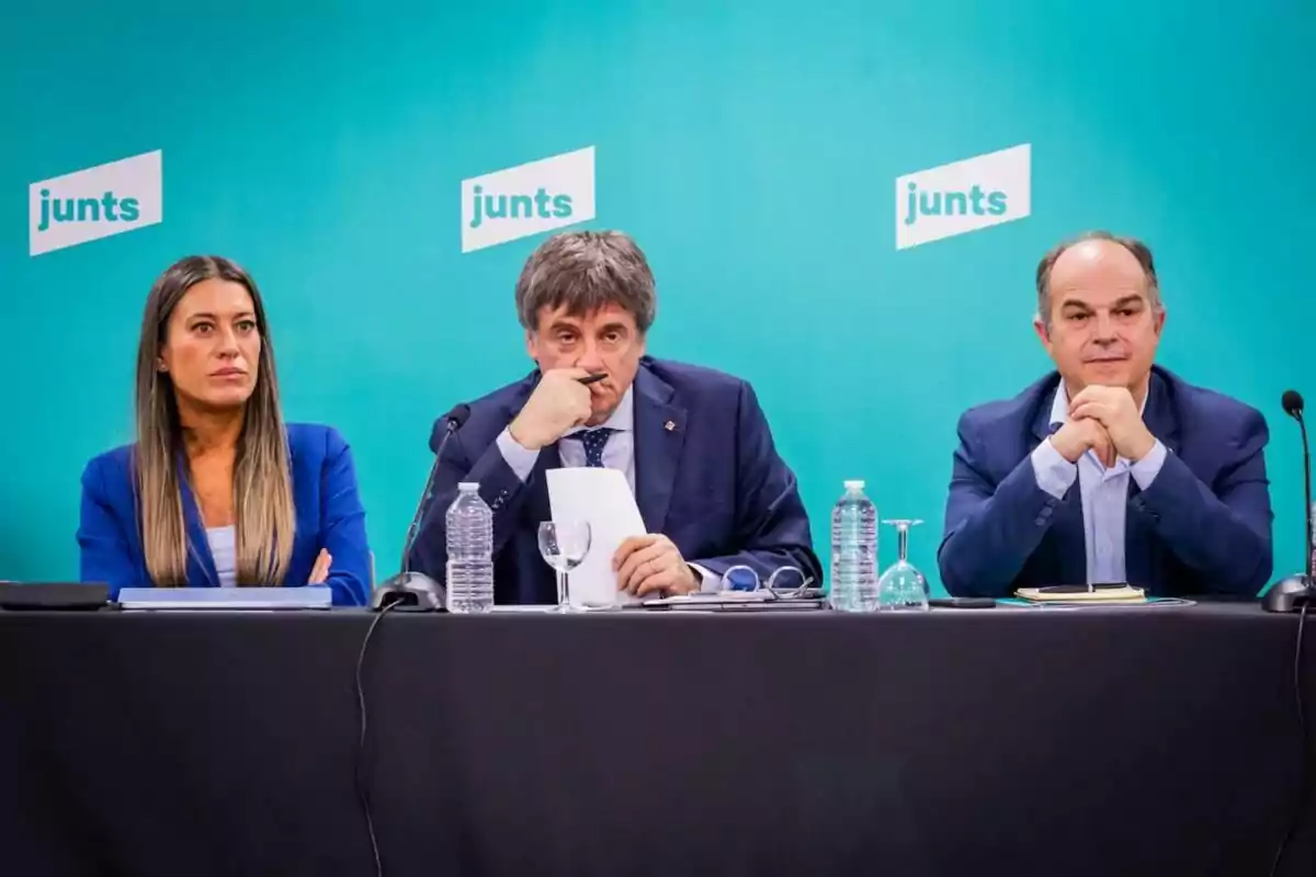 Tres personas sentadas en una mesa durante una conferencia de prensa con fondo turquesa y el logo de junts