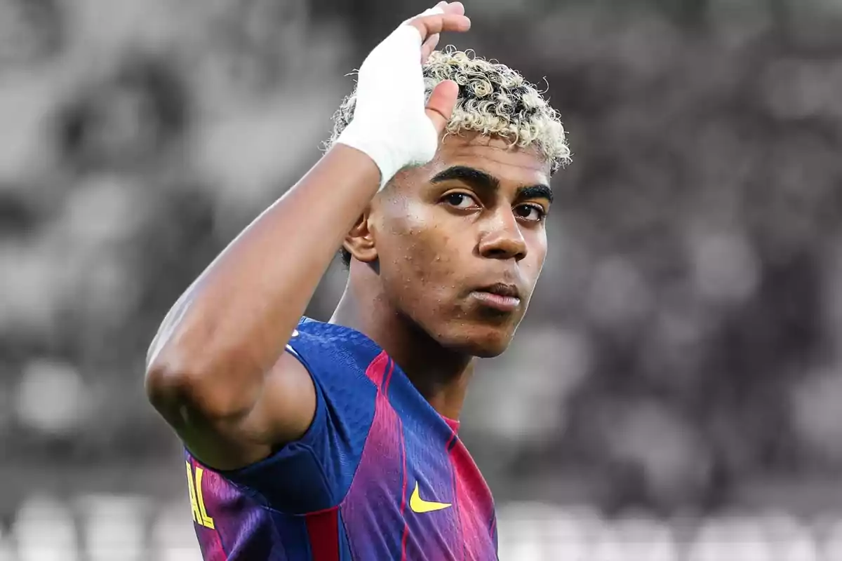 Jove futbolista amb els cabells arrissats i tenyits de ros, amb un embenat blanc a la mà i vestit amb un uniforme blau i vermell, aixeca la mà en senyal de salutació.