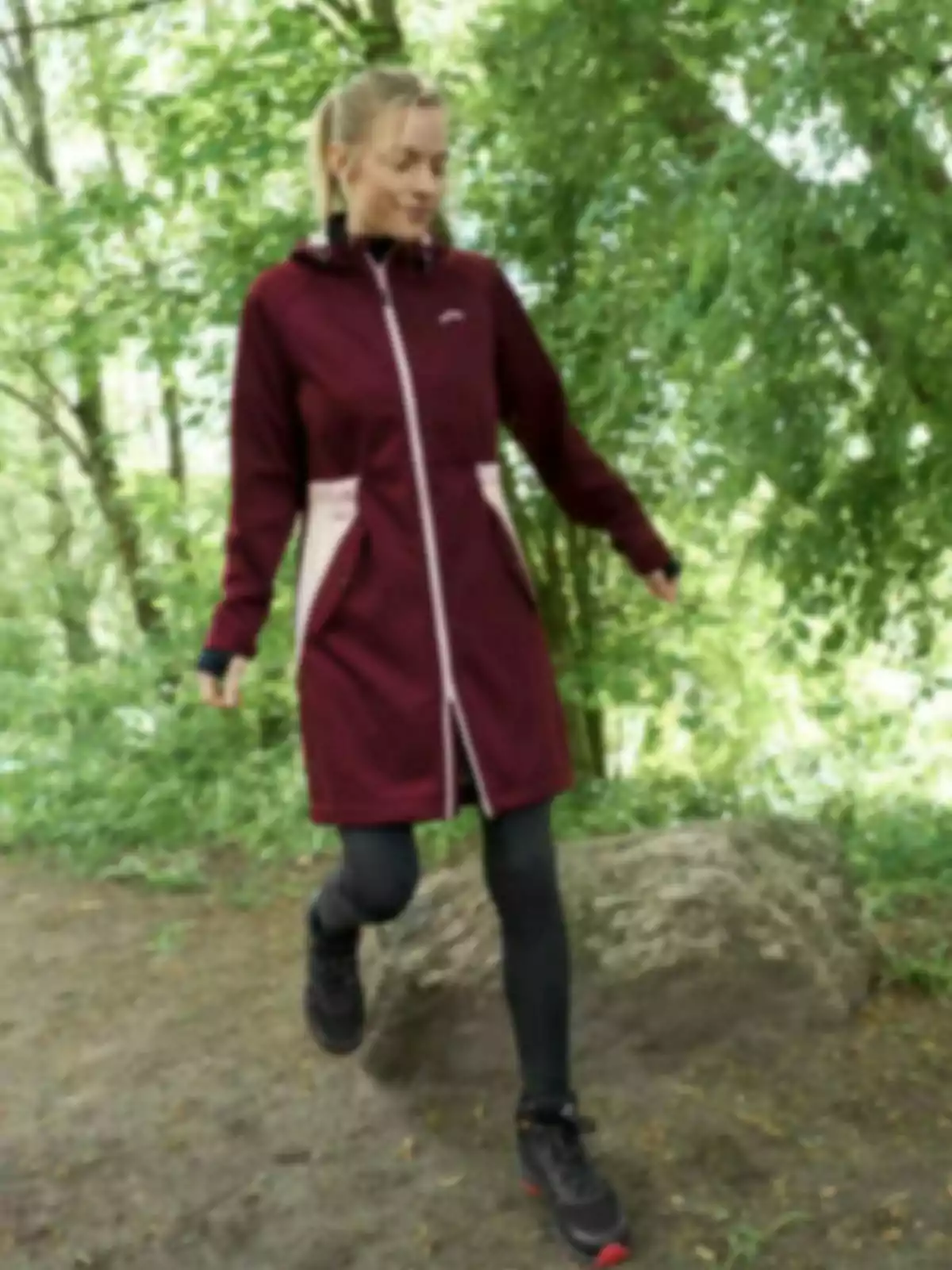 Mujer con abrigo burdeos y mallas negras caminando en un entorno natural lleno de árboles y vegetación