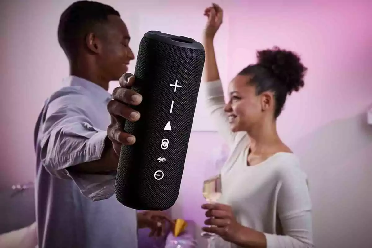 Un home sosté un altaveu portàtil mentre una dona somriu i balla en un ambient il·luminat amb llum tènue. Un home sosté un altaveu portàtil mentre una dona somriu i balla en un ambient il·luminat amb llum tènue.