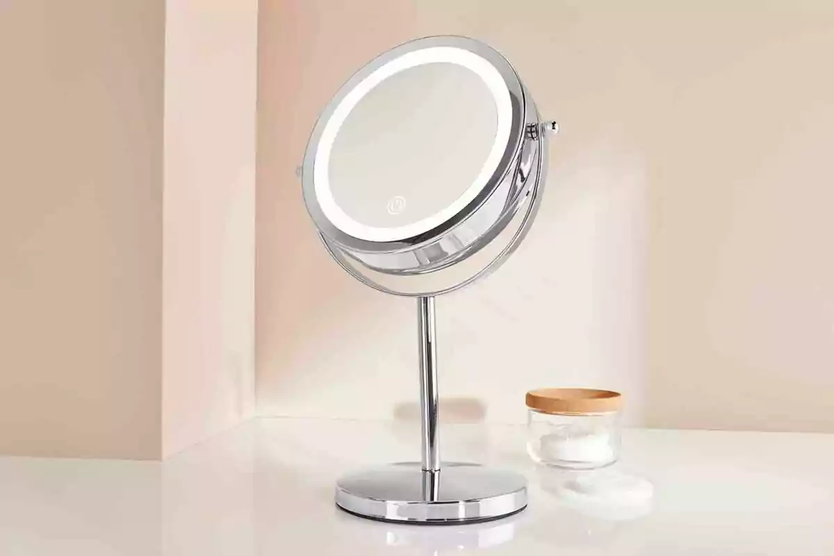 Espejo de aumento con luz LED sobre una superficie blanca junto a un frasco pequeño con tapa de madera y algunos discos de algodón