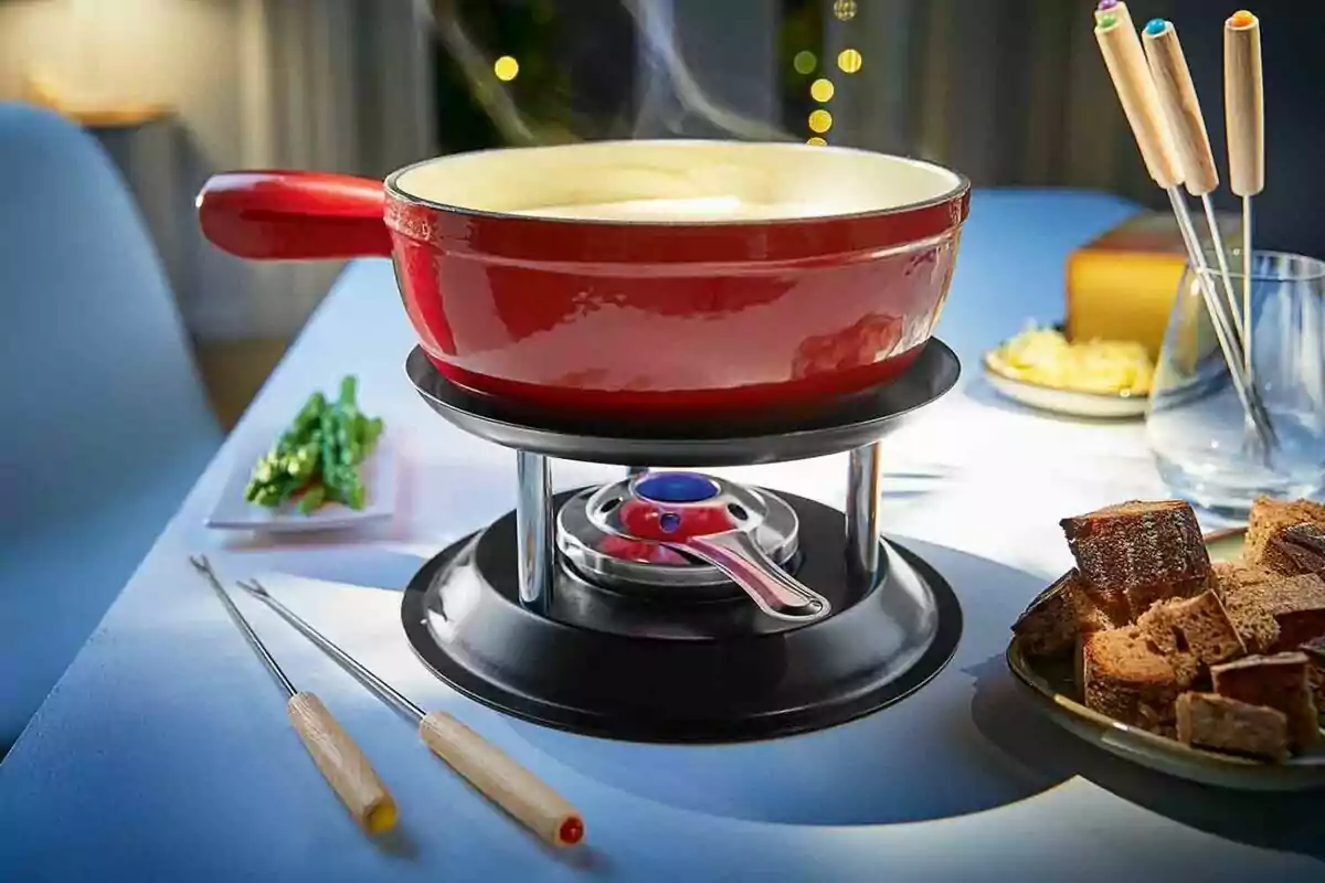 Olla de fondue roja sobre una mesa con pan, verduras y tenedores especiales alrededor